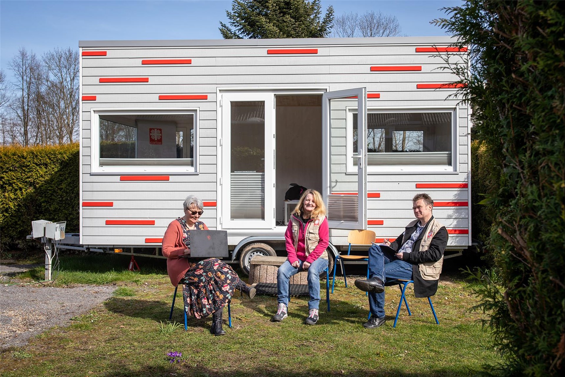 Drei Personen sitzen in der Sonne vor einem Tinyhouse