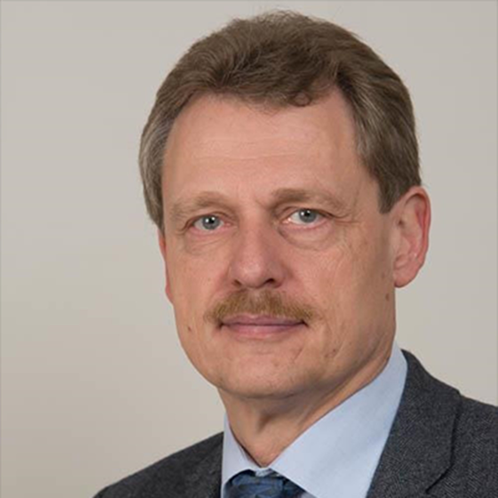 Mann mit blonden Haaren und Schnäuzer, der ein blaues Hemd und eine graue Anzugjacke trägt